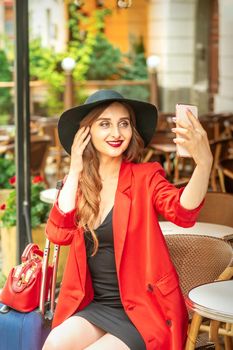 Tourist young caucasian woman in a red jacket and black hat with suitcase takes a selfie on a city street