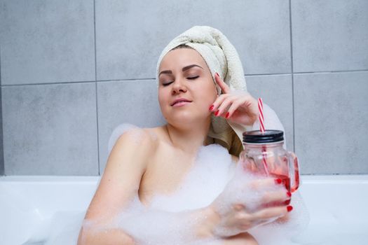Beautiful young woman with a towel on her head drinking a cocktail takes a bath at home. Relax after a hard day. The spa is a relaxing procedure.