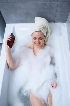 Beautiful young woman listening to music and singing while taking a bath at home. Relax after a hard day. Spa relaxing procedure.