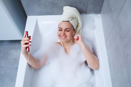 Beautiful young woman listening to music and singing while taking a bath at home. Relax after a hard day. Spa relaxing procedure.