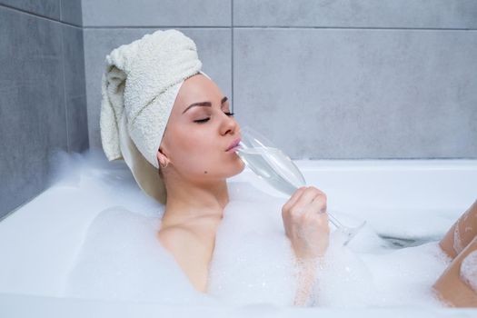 Girl woman with a white towel on her head lies with a glass of champagne in a white bathtub. In her hands a lot of pee soap. Full whirlpool foam. Relax after a hard day. Spa relaxing procedure