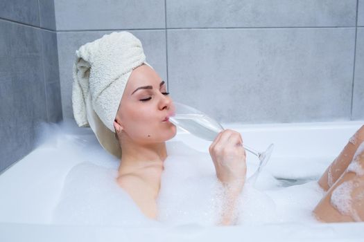 Girl woman with a white towel on her head lies with a glass of champagne in a white bathtub. In her hands a lot of pee soap. Full whirlpool foam. Relax after a hard day. Spa relaxing procedure