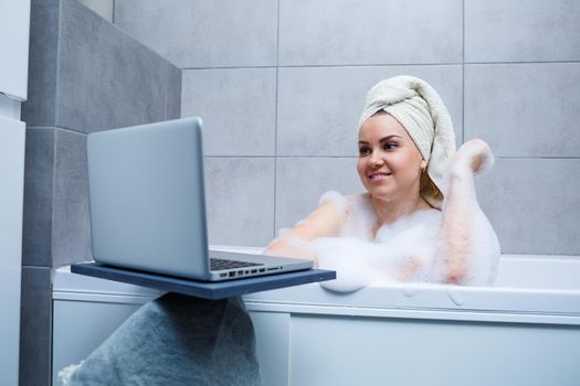 A young woman with a towel on her head is watching a movie on a laptop while sitting in a bath in a beauty salon. Relax in the bathroom without linen. Body care and relaxation concept.