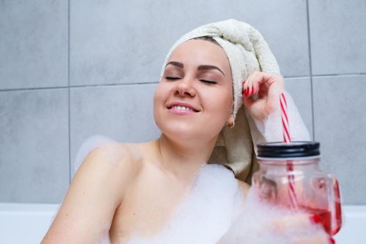 Beautiful young woman with a towel on her head, drinking a cocktail, takes a bath at home. Relax after a hard day.