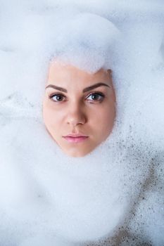 Closeup portrait of a girl in a bathtub and white foam around her face. Different emotions, grimaces, woman smile