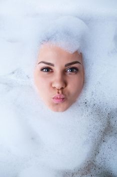 Closeup portrait of a girl in a bathtub and white foam around her face. Different emotions, grimaces, woman smile