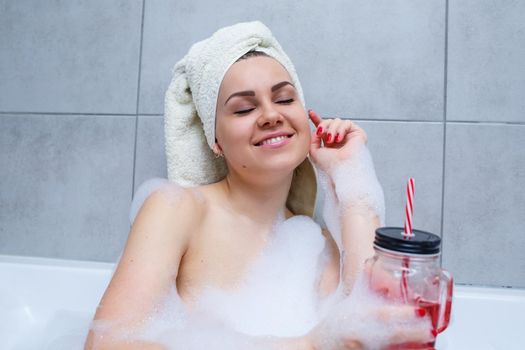 Beautiful young woman with a towel on her head drinking a cocktail takes a bath at home. Relax after a hard day. The spa is a relaxing procedure.