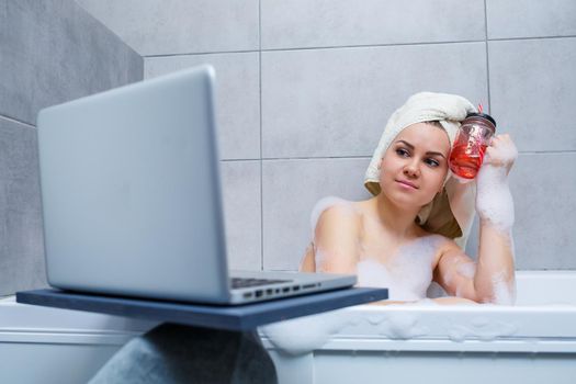 A young woman with a towel on her head drinks juice watching a movie on a laptop while sitting in a bath in a beauty salon. Relax in the bathroom without linen. Body care and relaxation concept.