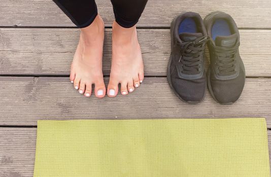 Close-up of bare feet on a wooden floor near a green sports mat, there are black sneakers nearby, in summer, near a pond in the park. Top view. Copy space