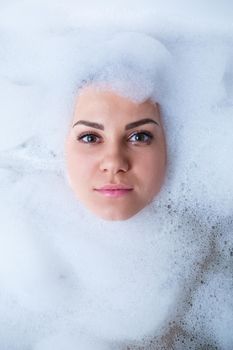 Closeup portrait of a girl in a bathtub and white foam around her face. Different emotions, grimaces, woman smile
