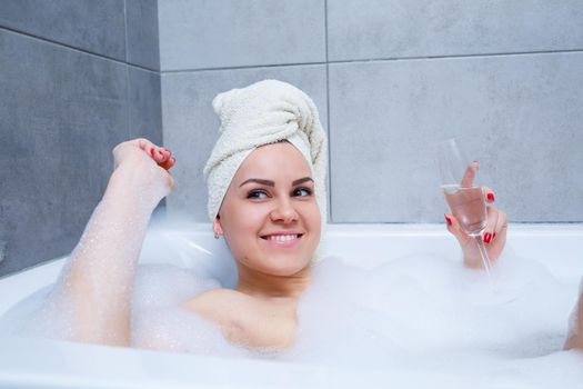 Girl woman with a white towel on her head lies with a glass of champagne in a white bathtub. In her hands a lot of pee soap. Full whirlpool foam. Relax after a hard day. Spa relaxing procedure