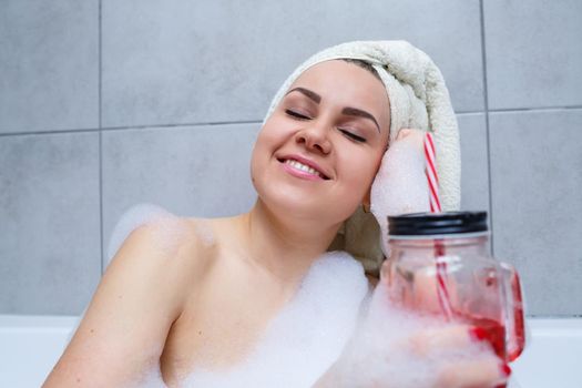 Beautiful young woman with a towel on her head drinking a cocktail takes a bath at home. Relax after a hard day. The spa is a relaxing procedure.