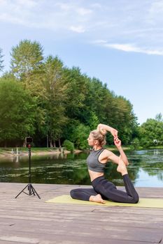 A slender woman in a gray top and leggings, on a wooden platform by a pond in the park in summer, does yoga, on a green sports mat, in front of her smartphone on a tripod.