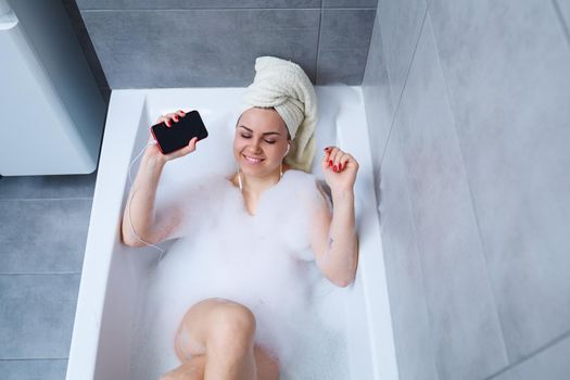 Beautiful young woman listening to music and singing while taking a bath at home. Relax after a hard day. Spa relaxing procedure.