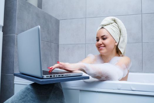 A young woman with a towel on her head is watching a movie on a laptop while sitting in a bath in a beauty salon. Relax in the bathroom without linen. Body care and relaxation concept.