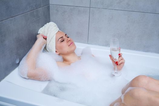 Girl woman with a white towel on her head lies with a glass of champagne in a white bathtub. In her hands a lot of pee soap. Full whirlpool foam. Relax after a hard day. Spa relaxing procedure