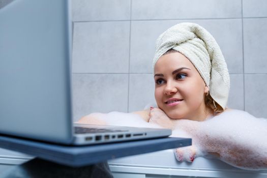 A young woman with a towel on her head is watching a movie on a laptop while sitting in a bath in a beauty salon. Relax in the bathroom without linen. Body care and relaxation concept.