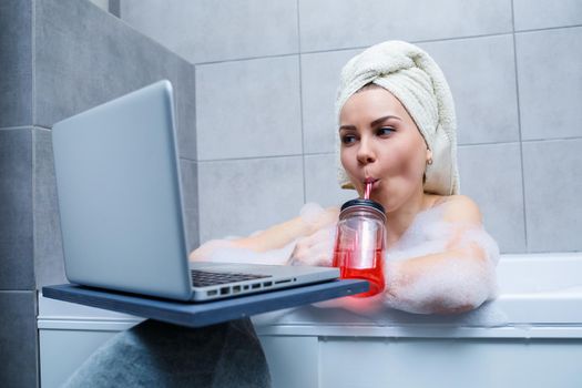 A young woman with a towel on her head drinks juice watching a movie on a laptop while sitting in a bath in a beauty salon. Relax in the bathroom without linen. Body care and relaxation concept.
