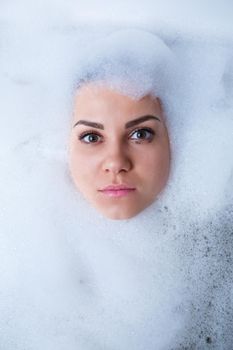 Closeup portrait of a girl in a bathtub and white foam around her face. Different emotions, grimaces, woman smile