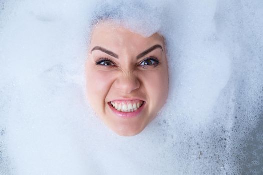 Closeup portrait of a girl in a bathtub and white foam around her face. Different emotions, grimaces, woman smile