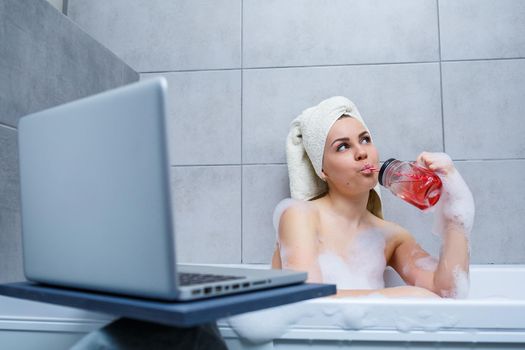 A young woman with a towel on her head drinks juice watching a movie on a laptop while sitting in a bath in a beauty salon. Relax in the bathroom without linen. Body care and relaxation concept.