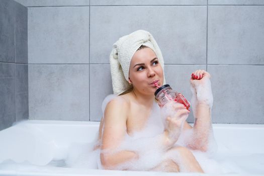 Beautiful young woman with a towel on her head drinking a cocktail takes a bath at home. Relax after a hard day. The spa is a relaxing procedure.