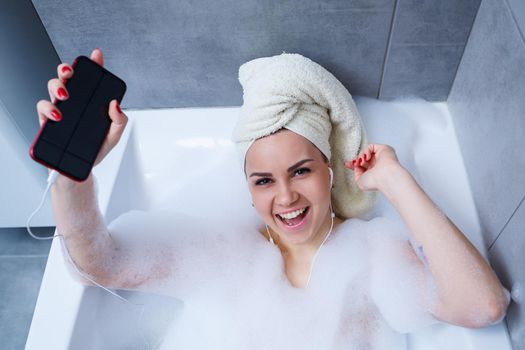 Beautiful young woman listening to music and singing while taking a bath at home. Relax after a hard day. Spa relaxing procedure.