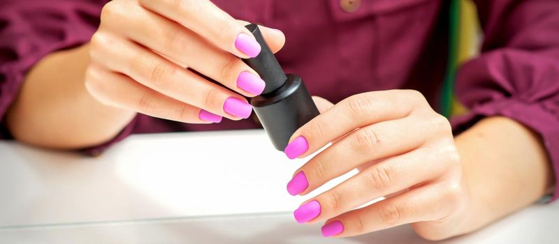 White woman holding nail polish black bottle with painted pink nails close up