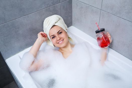 Beautiful young woman with a towel on her head drinking a cocktail takes a bath at home. Relax after a hard day. The spa is a relaxing procedure.