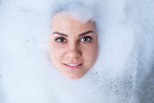 Closeup portrait of a girl in a bathtub and white foam around her face. Different emotions, grimaces, woman smile