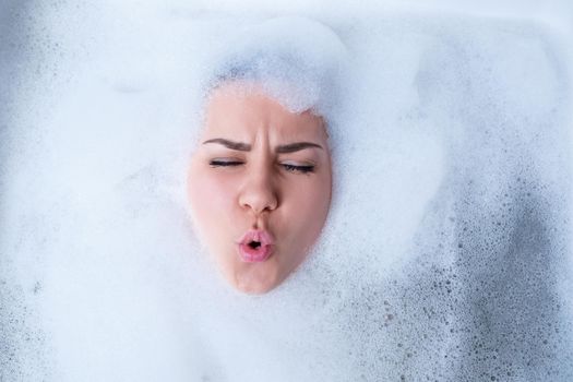 Closeup portrait of a girl in a bathtub and white foam around her face. Different emotions, grimaces, woman smile