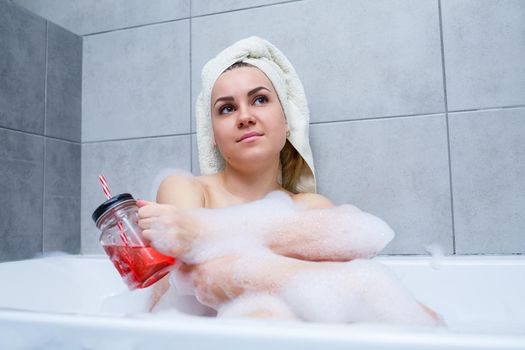 Beautiful young woman with a towel on her head drinking a cocktail takes a bath at home. Relax after a hard day. The spa is a relaxing procedure.
