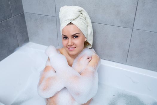 Girl woman with a white towel on her head lies in the bathroom in a white bath. Foam for around. Relax after a hard day. Spa relaxing treatment