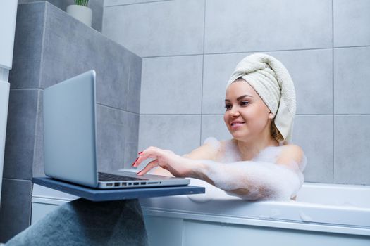 A young woman with a towel on her head is watching a movie on a laptop while sitting in a bath in a beauty salon. Relax in the bathroom without linen. Body care and relaxation concept.