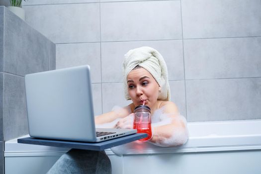 A young woman with a towel on her head drinks juice watching a movie on a laptop while sitting in a bath in a beauty salon. Relax in the bathroom without linen. Body care and relaxation concept.