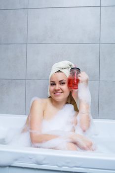 Beautiful young woman with a towel on her head, drinking a cocktail, takes a bath at home. Relax after a hard day.