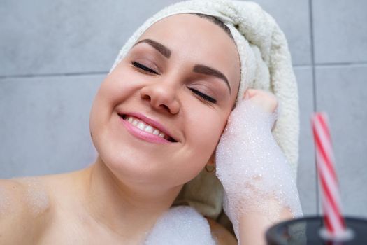Beautiful young woman with a towel on her head drinking a cocktail takes a bath at home. Relax after a hard day. The spa is a relaxing procedure.