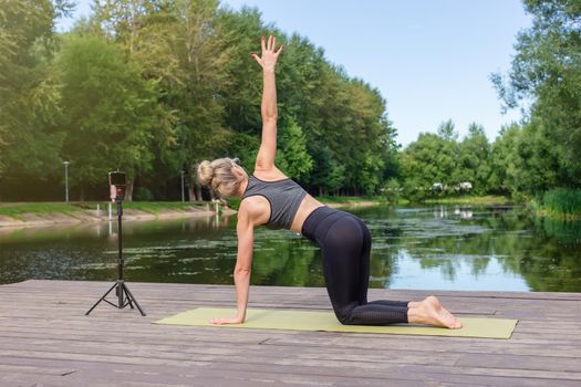 A slender woman in a gray top and leggings, on a wooden platform by a pond in the park in summer, does yoga, performs exercises, on a green sports mat, in front of her smartphone on a tripod.