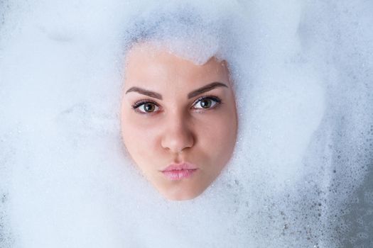 Closeup portrait of a girl in a bathtub and white foam around her face. Different emotions, grimaces, woman smile