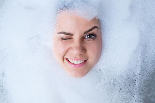 Closeup portrait of a girl in a bathtub and white foam around her face. Different emotions, grimaces, woman smile