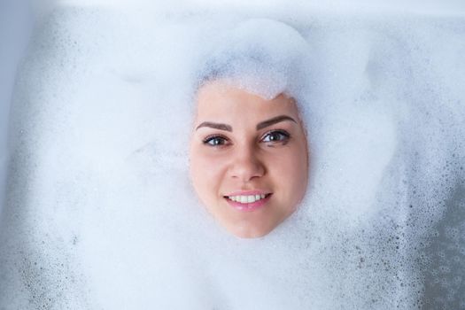 Closeup portrait of a girl in a bathtub and white foam around her face. Different emotions, grimaces, woman smile