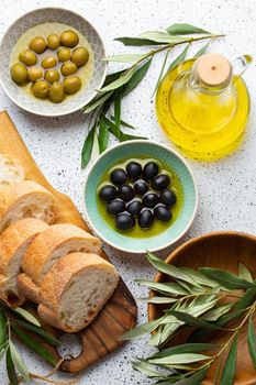 Green and black olives with olive oil in a glass bottle, olive tree sprigs and cut fresh ciabatta bread on wooden cutting board. White rustic background, mediterranean food concept, top view