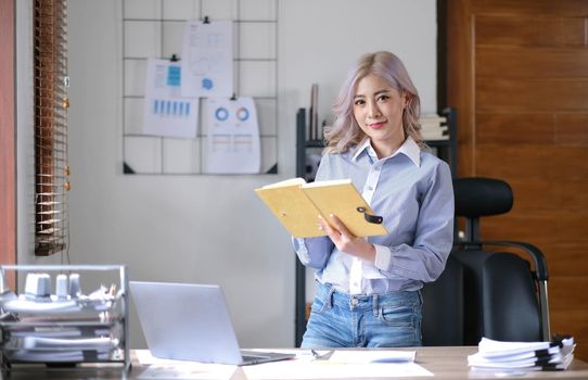 Charming young Asian businesswoman with a smile standing holding book at the office. Looking at camera..