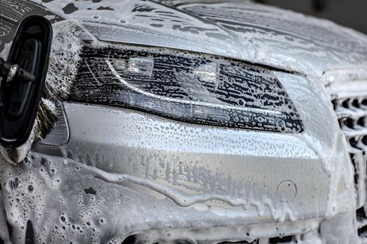 Front light of silver car washed in manual carwash, brush leaving strokes in shampoo and foam.