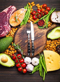 Knife and fork over wooden cutting board and ketogenic low carbs ingredients for healthy eating concept and weight loss, top view. Keto foods: meat, fish, avocado, cheese, vegetables, nuts
