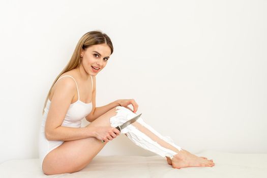 Beautiful caucasian woman shaving her legs with a knife with shaving foam on white background. Depilation and epilation concept