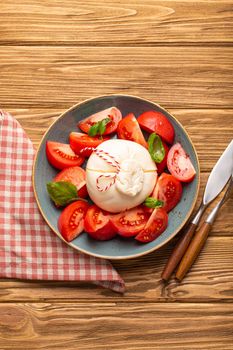 Italian salad with fresh tomatoes, burrata cheese, basil and olive oil served on ceramic plate on rustic wooden background from above, healthy snack or lunch