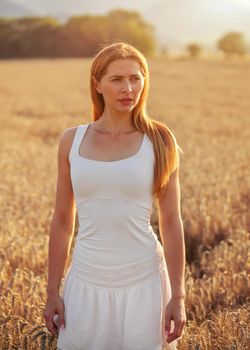 Young sporty woman in white dress, long brown hair, looking into distance, sunset lit wheat field behind her.