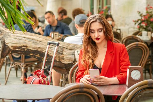 Tourist young caucasian white woman in a red jacket looking at smartphone sitting at the table in cafe outdoors