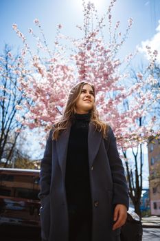 Sakura branches with flowers on a tree on the city streets. Stylish woman girl stands in the street with blooming sakura. Modern fashionable girl outdoors. Sakura tree blossoms.